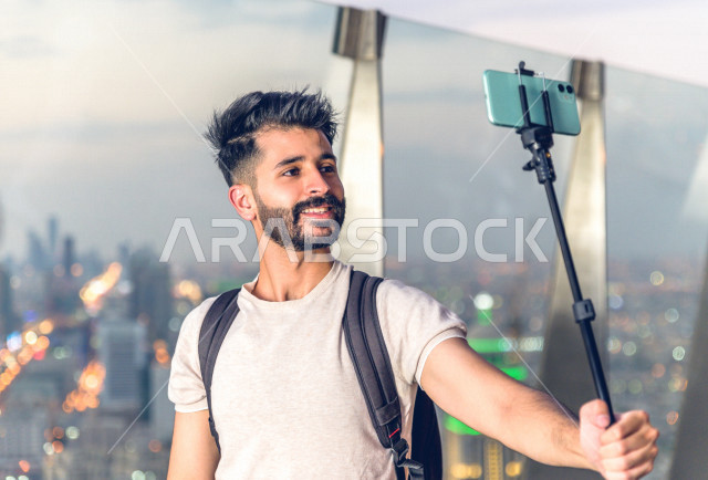 شاب سعودي سائح ، يلتقط صورة سلفي باطلالة ابراج الرياض من الخلف ، مصور خليجي يحمل هاتفه المحمول ، يلتقط صور سلفي 