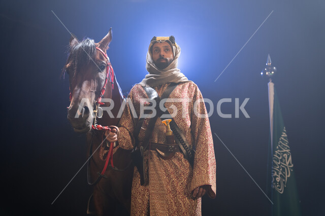 الوقوف بجانب علم السعودية باعتزاز، اليوم الوطني23 سبتمبر،  ارتداء أزياء المناسبات والأعياد الوطنية، ذكرى تأسيس الدولة السعودية الأولى 1727 م، يوم العلم السعودي 11 مارس، رجل خليجي يرتدي الدقلة والغترة يقف مع خيله العربي الأصيل، خلفية ضبابية مع إضاءة بيضاء.