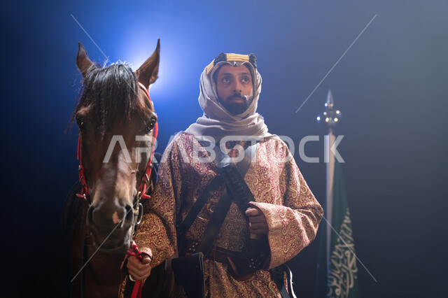 الوقوف بجانب علم السعودية باعتزاز، اليوم الوطني23 سبتمبر، المناسبات والأعياد الوطنية، ذكرى تأسيس الدولة السعودية الأولى 1727 م، يوم العلم السعودي 11 مارس، صورة مقربة لرجل خليجي يرتدي الدقلة والغترة يقف مع خيله العربي الأصيل، خلفية ضبابية مع إضاءة بيضاء. 