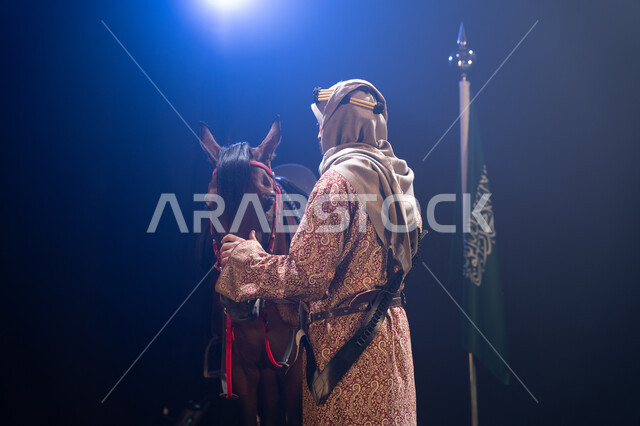 يوم العلم السعودي 11 مارس، الوقوف بجانب علم السعودية باعتزاز، اليوم الوطني23 سبتمبر، المناسبات والأعياد الوطنية، ذكرى تأسيس الدولة السعودية الأولى 1727 م، صورة جانبية لرجل خليجي يرتدي الدقلة والغترة يقف مع خيله العربي الأصيل، خلفية ضبابية مع إضاءة بيضاء. 