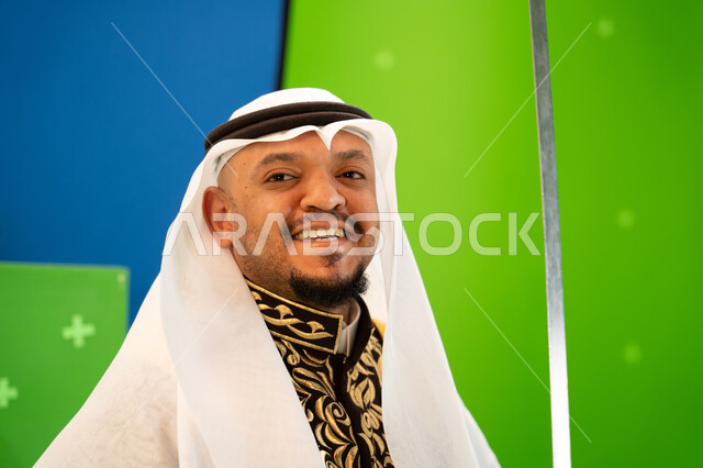 النظر إلى الكاميرا بإيماءات السعادة ، العيد الوطني السعودي 23 سبتمبر، الرقصات والفعاليات الشعبية و الأهازيج في العرضة النجدية في المناسبات الوطنية، صورة مقربة لرجل عربي سعودي يرتدي الغترة والدقلة يرقص بالسيف، خلفية خضراء وزرقاء ،كروما