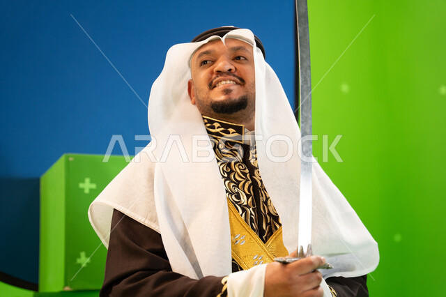 الاحتفال بذكرى اليوم الوطني السعودي بإيماءات السعادة ،العيد الوطني السعودي 23 سبتمبر، الرقصات والفعاليات الشعبية و الأهازيج في العرضة النجدية في المناسبات الوطنية، صورة مقربة لرجل عربي سعودي يرتدي الغترة والدقلة يرقص بالسيف، خلفية خضراء وزرقاء ،كروما