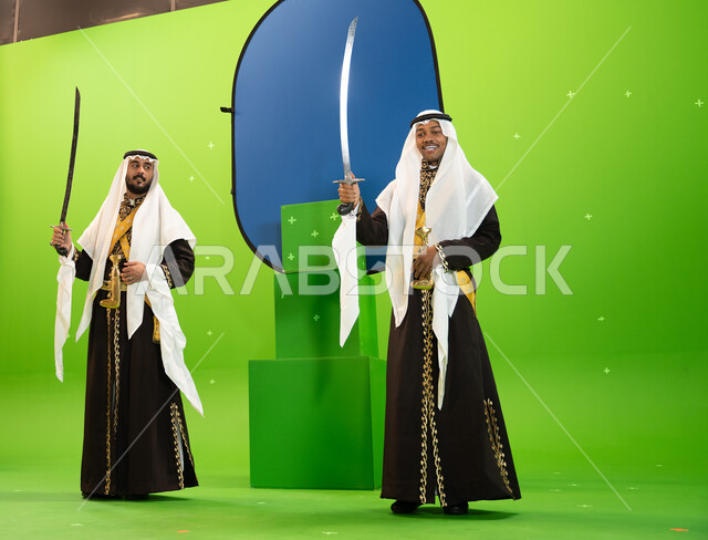 الاحتفال بذكرى اليوم الوطني السعودي 23 سبتمبر ، إظهار إرث المنطقة الجنوبية, استخدام السيوف في الرقص الشعبي، رجلان عربيان سعوديان من فرقة العرضة النجديه يرتديان الغترة والدقله، صورة الجسم بالطول الكامل، خلفيه خضراء و زرقاء ،كروما