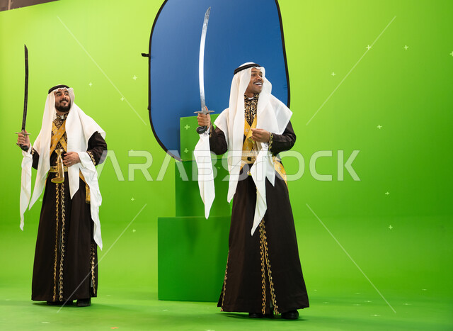 الاحتفال بذكرى تأسيس الدولة السعودية الاولى 22 شباط ، إظهار إرث المنطقة الجنوبية, استخدام السيوف في الرقص الشعبي، رجلان عربيان سعوديان من فرقة العرضة النجديه يرتديان الغترة والدقله، صورة الجسم بالطول الكامل، خلفيه خضراء و زرقاء ،كروما