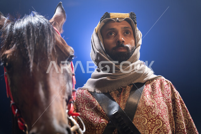 توارث حب الجياد من الأجداد، الوقوف بفخر واعتزاز والنظر إلى شيء ما، ارتداء أزياء المناسبات والأعياد الوطنية، ذكرى تأسيس الدولة السعودية الأولى 1727 م، صورة مقربة لرجل خليجي يرتدي الدقلة والغترة يقف مع خيله العربي الأصيل، خلفية ضبابية مع إضاءة بيضاء. 