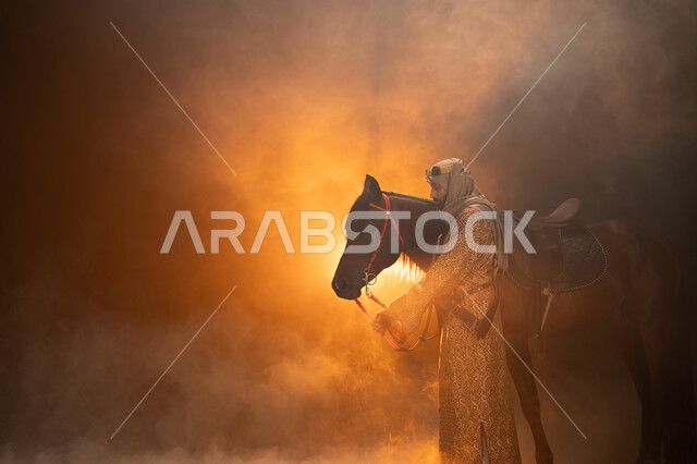 يوم التأسيس السعودي 22 فبراير، حب الفروسية والشغف في تربية الجياد، ارتداء أزياء المناسبات والأعياد الوطنية، ذكرى تأسيس الدولة السعودية الأولى 1727 م، صورة جانبية لرجل خليجي يرتدي الدقلة والغترة يقف مع خيله العربي الأصيل، خلفية ضبابية مع إضاءة صفراء. 