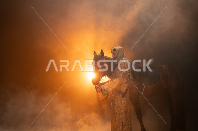 يوم التأسيس السعودي 22 فبراير، حب الفروسية والشغف في تربية الجياد، ارتداء أزياء المناسبات والأعياد الوطنية، ذكرى تأسيس الدولة السعودية الأولى 1727 م، صورة جانبية لرجل خليجي يرتدي الدقلة والغترة يقف مع خيله العربي الأصيل، خلفية ضبابية مع إضاءة صفراء. 