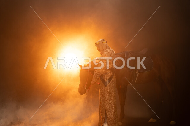 يوم التأسيس السعودي 22 فبراير، حب الفروسية والشغف في تربية الجياد، ارتداء أزياء المناسبات والأعياد الوطنية، ذكرى تأسيس الدولة السعودية الأولى 1727 م، صورة جانبية لرجل خليجي يرتدي الدقلة والغترة يقف مع خيله العربي الأصيل، خلفية ضبابية مع إضاءة صفراء. 