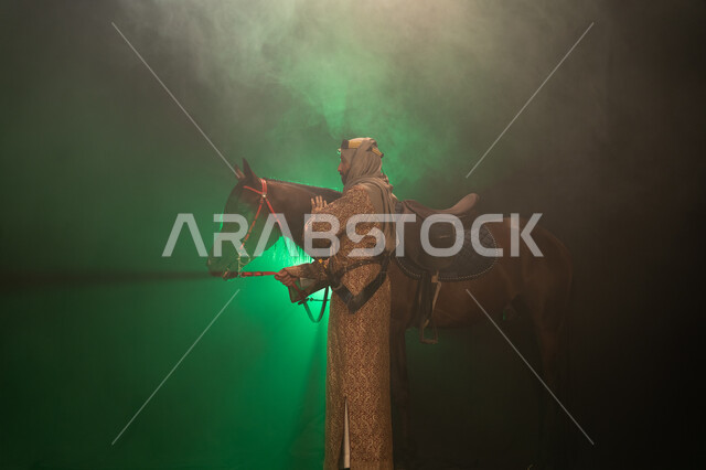 ارتداء أزياء المناسبات والأعياد الوطنية، نشر حب الخيل بين أبناء الوطن، ذكرى تأسيس الدولة السعودية الأولى 1727 م، يوم التأسيس السعودي 22 فبراير، صورة جانبية لرجل خليجي يرتدي الدقلة والغترة يداعب خيله العربي الأصيل، خلفية ضبابية مع إضاءة خضراء. 