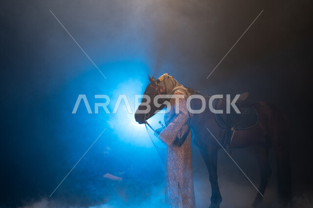 نشر حب الخيل بين أبناء الوطن، ذكرى تأسيس الدولة السعودية الأولى 1727 م، يوم التأسيس السعودي 22 فبراير، ارتداء أزياء المناسبات والأعياد الوطنية، صورة جانبية لرجل خليجي يرتدي الدقلة والغترة يداعب خيله العربي الأصيل، خلفية ضبابية مع إضاءة زرقاء. 
