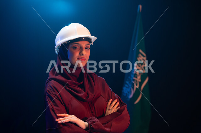 تطوير القطاع الهندسي ومجال الاعمار في السعودية، يوم العلم 11 مارس، الاحتفال بذكرى اليوم الوطني السعودي 23 سبتمبر، صورة مقربة لمهندسة عربية خليجية سعودية محجبة ترتدي خوذة الحماية تقف بثقة وأيدي متقاطعة جانب علم السعودية، خلفية سوداء مع اضاءة بيضاء