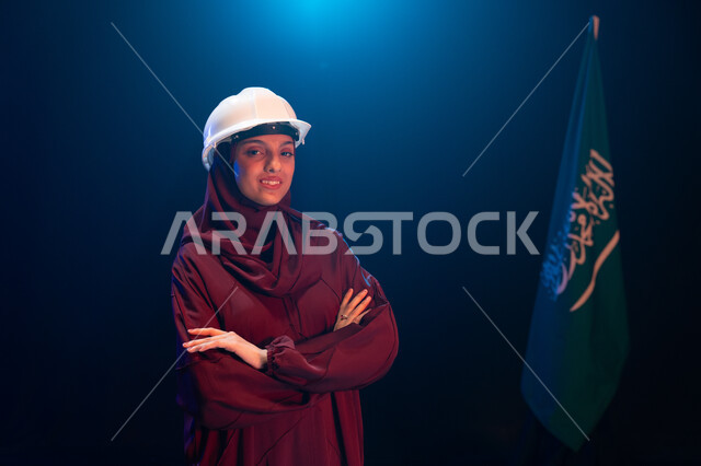 يوم العلم 11 مارس، تنمية القطاع الهندسي ومجال الاعمار في السعودية، الاحتفال بذكرى اليوم الوطني السعودي 23 سبتمبر، صورة مقربة لمهندسة عربية خليجية سعودية مبتمسة ترتدي الحجاب وخوذة الحماية تقف بثقة وأيدي متقاطعة جانب علم السعودية، خلفية سوداء مع اضاءة بيضاء