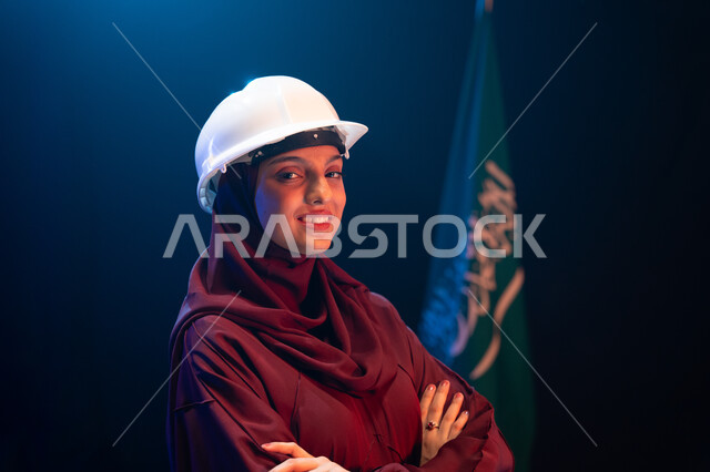 الاحتفال بيوم العلم 11 مارس، تنمية القطاع الهندسي ومجال الاعمار في السعودية، ذكرى اليوم الوطني السعودي 23 سبتمبر، صورة مقربة لمهندسة عربية خليجية سعودية مبتمسة ترتدي الحجاب وخوذة الحماية تقف بثقة وأيدي متقاطعة جانب علم السعودية، خلفية سوداء مع اضاءة بيضاء