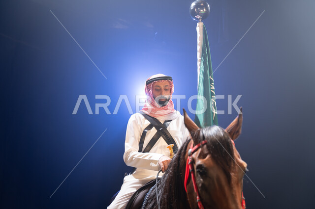 إحياء ذكرى العيد الوطني، الوقوف بفخر بجانب علم السعودية، خلفية ضبابية مع إضاءة بيضاء، الاحتفال مع الخيل بذكرى يوم العلم السعودي11 مارس، صورة مقربة لرجل عربي خليجي سعودي يرتدي الشماغ والثوب التقليدي يمتطي فرسه البني العربي الأصيل. 