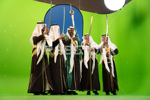 الرقص الشعبي في المناسبات الوطنيه والاعياد، استخدام السيف بحركات منتظمه في العرضة السعوديه ،ذكرى اليوم الوطني السعودي في 23 سبتمبر، ايماءات الضحك والسرور ، الغناء والقاء القصائد الحماسيه والشعر السعودي، صورة الجسم بالطول الكامل، خلفية خضراء وزرقاء، كروما
