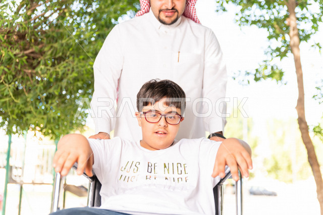 ولد جميل سعوي خليجي مصاب بملازمة داون يقضي وقته في نزهه مع والده  بالمنتزه ، طفل مقعد على كرسي متحرك يستمتع بالهواء الطلق ، أصحاب الهمم ، ذوي الاحتياجات الخاصة ، اعاقة حركية