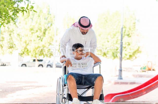 A Gulf boy with Down syndrome spends his time outing with his father in the park, a wheelchair-bound child enjoying the outdoors, people of determination, special needs, mobility impairment
