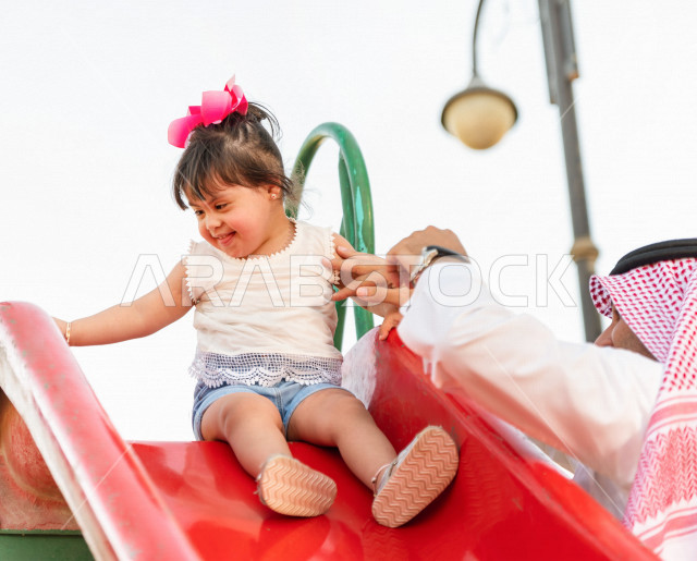 A beautiful girl with Down syndrome spends her time outing with her father in the park, a Saudi Gulf father plays with his daughter in the open air, slides and swings in the park on a sunny day  People of determination, people with special needs