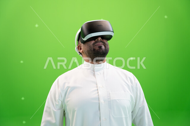 النظر إلى الأعلى بايماءات الاندماج والتفاعل، استخدام التقنيات الحديثة المتطورة في مشاهدة الأفلام و محاكاة العالم الخيالي ، بورتريه مقرب لرجل عربي خليجي سعودي يرتدي الزي التقليدي يلبس نظارة الواقع الإفتراضي ثلاثية الأبعاد، خلفية خضراء، كروما