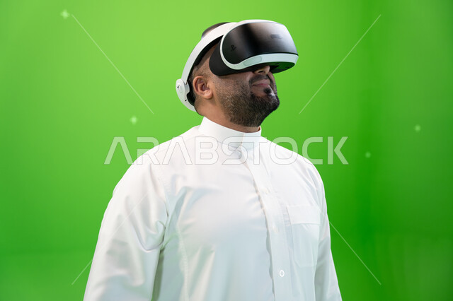Looking up with gestures of interaction and integration, the use of advanced modern technologies in watching movies and simulating the imaginary world, a close-up portrait of a Saudi Arabian Gulf man in traditional dress wearing 3D virtual reality glasses