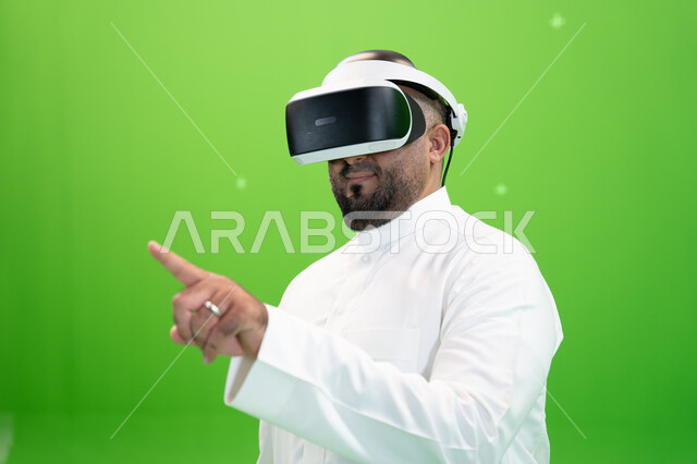 اشارات النقر على شاشه هولوجرامية ، بورتريه مقرب من الجانب لرجل عربي خليجي سعودي يرتدي الزي التقليدي يلبس نظارة الواقع الإفتراضي ثلاثية الأبعاد، خلفية خضراء، كروما