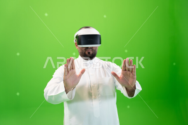 اشارات يدين تدل على اللمس والاحساس و دفع عنصر هولوجرامي ، بورتريه مقرب  لرجل عربي خليجي سعودي يرتدي الزي التقليدي يلبس نظارة الواقع الإفتراضي ثلاثية الأبعاد، خلفية خضراء، كروما
