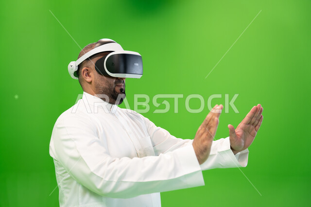 ايماءات يدين تدل على اللمس والاحساس و دفع عنصر هولوجرامي ، بورتريه مقرب من الجانب لرجل عربي خليجي سعودي يرتدي الزي التقليدي يلبس نظارة الواقع الإفتراضي ثلاثية الأبعاد، خلفية خضراء، كروما