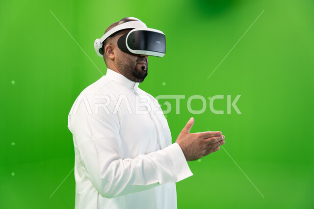 استخدام  التقنيات المتطورة  الحديثة في مشاهدة الأفلام و محاكاة العالم الخيالي ، بورتريه مقرب لرجل عربي خليجي سعودي يرتدي الزي التقليدي يلبس نظارة الواقع الإفتراضي ثلاثية الأبعاد، ايماءات التفاعل والاندماج ، اشارات التحيه والسلام, خلفية خضراء، كروما