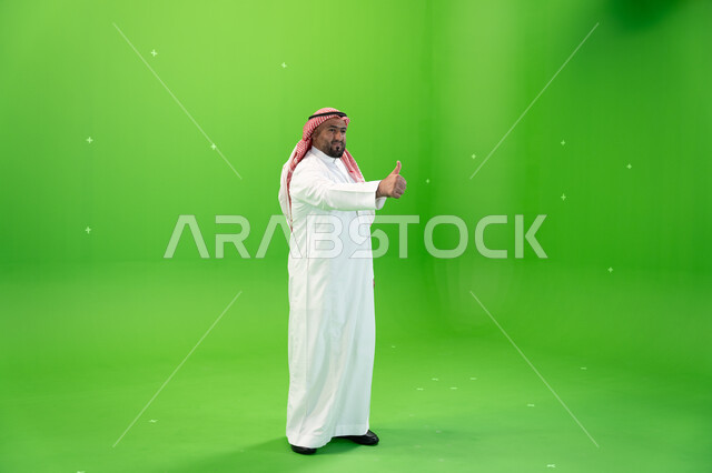 اشارات يدين تدل على الرضى والاعجاب وعلامة الجودة، بورتريه لرجل عربي خليجي سعودي يرتدي الثوب والشماغ التقليدي يقوم برفع الابهام الى الاعلى، صورة الجسم بالطول الكامل،  خلفية خضراء، كروما