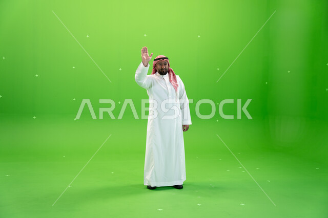 ايماءات السلام و إلقاء التحية  و السعادة بالترحيب، بورتريه لرجل عربي خليجي سعودي يرتدي الثوب والشماغ التقليدي يقوم برفع يده اليمنى للأعلى ، صورة الجسم بالطول الكامل،  خلفية خضراء، كروما