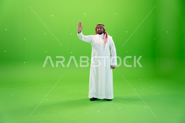 ايماءات إلقاء التحية والسلام و السعادة بالترحيب، بورتريه لرجل عربي خليجي سعودي يرتدي الثوب والشماغ التقليدي يقوم برفع يده اليمنى للأعلى ، صورة الجسم بالطول الكامل،  خلفية خضراء، كروما