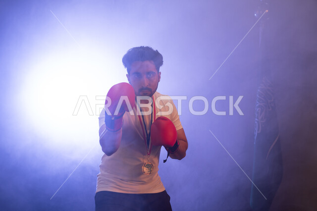نشاط بدني للحفاظ على الصحة واللياقة الجسدية، المنتخب الوطني السعودي للمصارعة، صورة مقربة لرجل عربي خليجي يرتدي قفازات الملاكمة والميدالية الذهبية يتدرب على الحركات القتالية، ممارسة رياضة الدفاع عن النفس ، تفريغ الطاقة السلبية، خلفية ضبابية مع إضاءة بيضاء.