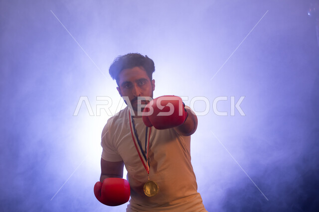 نشاط بدني للحفاظ على الصحة واللياقة الجسدية، المنتخب الوطني السعودي للمصارعة، صورة مقربة لرجل عربي خليجي يرتدي قفازات الملاكمة والميدالية الذهبية يتدرب على الحركات القتالية، ممارسة رياضة الدفاع عن النفس ، تفريغ الطاقة السلبية، خلفية ضبابية مع إضاءة بيضاء.