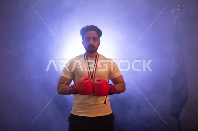 ممارسة رياضة الدفاع عن النفس، المنتخب الوطني السعودي للمصارعة، نشاط بدني للحفاظ على الصحة واللياقة الجسدية، رجل عربي خليجي سعودي يرتدي قفازات الملاكمة والميدالية الذهبية، التدرب على الحركات القتالية، تفريغ الطاقة السلبية، خلفية ضبابية مع إضاءة بيضاء.
