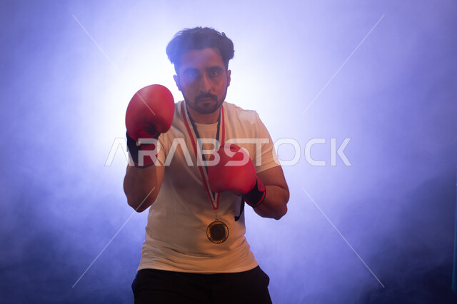 ممارسة رياضة الدفاع عن النفس، المنتخب الوطني السعودي للمصارعة، نشاط بدني للحفاظ على الصحة واللياقة الجسدية، رجل عربي خليجي سعودي يرتدي قفازات الملاكمة والميدالية الذهبية، التدرب على الحركات القتالية، تفريغ الطاقة السلبية، خلفية ضبابية مع إضاءة بيضاء.