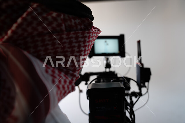 The talent and professionalism of photography, practicing the hobby of taking pictures with high-resolution cameras, a close-up of a Saudi Arabian Gulf man wearing the traditional dress and the shemagh holding the camera, taking souvenir photos, a white b
