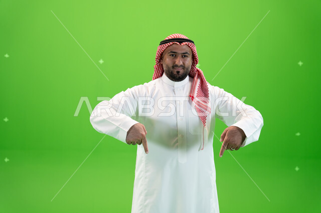حركات تدل على هذا المكان (هنا), في هذه اللحظه (الآن), إيماءات يدين تشير الى موقع معين, بورتريه  لرجل عربي خليجي سعودي يرتدي الثوب والشماغ التقليدي يشير بأصابعه نحو الأسفل وينظر إلى الكاميرا، خلفية خضراء، كروما