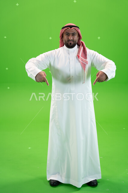  إيماءات يدين تشير الى موقع معين, في هذه اللحظه (الآن), بورتريه لرجل عربي خليجي سعودي يرتدي الثوب والشماغ التقليدي يشير بأصابعه نحو الأسفل بإيماءات السعاده، اشارات تدل على هذا المكان (هنا), صورة الجسم بالطول الكامل، خلفية خضراء، كروما