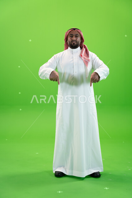 في هذه اللحظه (الآن), إيماءات يدين تشير الى موقع معين, بورتريه  لرجل عربي خليجي سعودي يرتدي الثوب والشماغ التقليدي يشير بأصابعه نحو الأسفل وينظر  إلى الكاميرا، حركات تدل على هذا المكان (هنا), صورة الجسم بالطول الكامل، خلفية خضراء، كروما