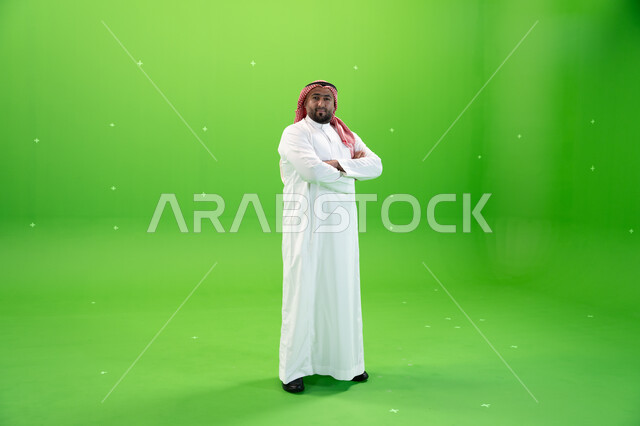 الثقه والاعتزاز بالنفس، بورتريه لرجل عربي خليجي سعودي يرتدي الثوب والشماغ التقليدي ينظر إلى الكاميرا، الوقوف بإيماءات يدين متقاطعة، صورة الجسم بالطول الكامل، خلفيه خضراء، كروما