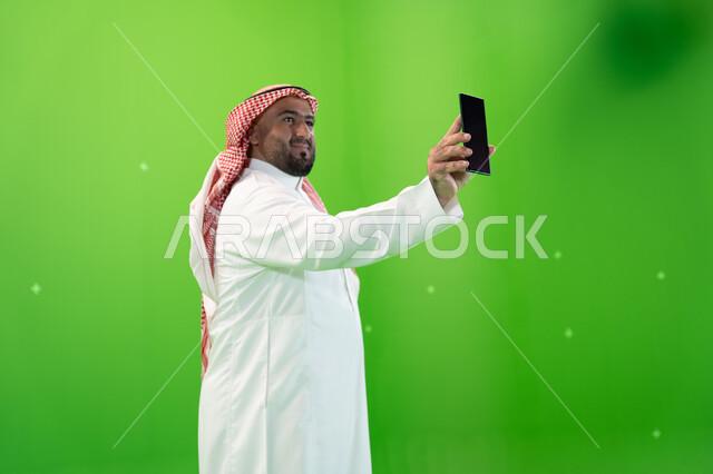 استخدام التقنيات الحديثه المتطورة، بورتريه لرجل عربي خليجي سعودي يرتدي الثوب والشماغ التقليدي و يقوم بعرض شاشه سوداء فارغه عبر الهاتف المحمول،  خلفية خضراء، كروما