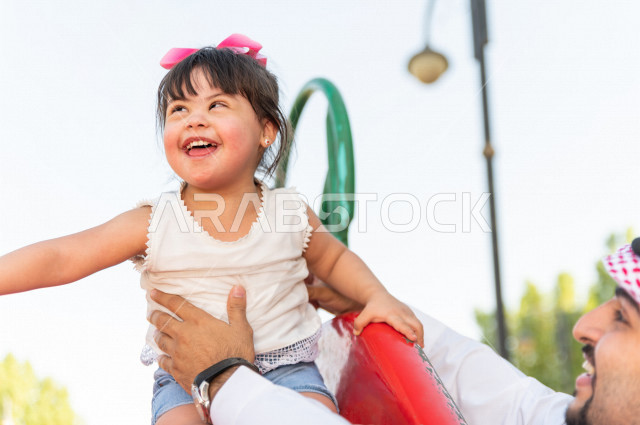 فتاه جميلة مصابة بملازمة داون تقضي وقتها في نزهه مع والدها بالمنتزه ، اب سعودي خليجي يلعب مع ابنته في الهواء الطلق ،تنزلق وتتأرجح في الحديقة في يوم مشمس  ، أصحاب الهمم ، ذوي الاحتياجات الخاصة