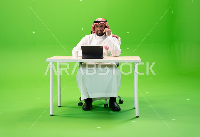 إجراء مكالمة هاتفية عبر الهاتف المحمول, بورتريه لموظف عربي سعودي خليجي يرتدي الثوب السعودي التقليدي والشماغ يقوم بالعمل داخل مكتبه, استخدام جهاز التابليت لإنجاز مهام العمل, إدارة شؤون الأعمال وتنظيمها, خلفية خضراء, كروما.