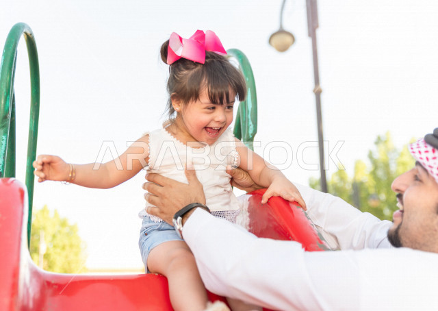 فتاه جميلة مصابة بملازمة داون تقضي وقتها في نزهه مع والدها بالمنتزه ، اب سعودي خليجي يلعب مع ابنته في الهواء الطلق ،تنزلق وتتأرجح في الحديقة في يوم مشمس  ، أصحاب الهمم ، ذوي الاحتياجات الخاصة