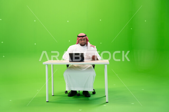 استخدام الأجهزة والتقنيات الحديثة, بورتريه لشاب عربي خليجي سعودي يرتدي الثوب السعودي التقليدي والشماغ يجلس في مكتبه ويقرأ التقرير, إنجاز مهام العمل عبر جهاز التابليت, متابعة وإدارة الأعمال الادارية, تصفح المواقع الالكترونية, خلفية خضراء, كروما.