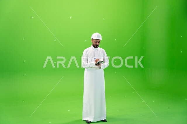 Recording and taking notes on building and construction, Saudi professions and jobs, working in the Saudi engineering sector, portrait of a Saudi Gulf Arab engineer wearing a safety hat standing at the work site, reconstruction with the arms of the nation