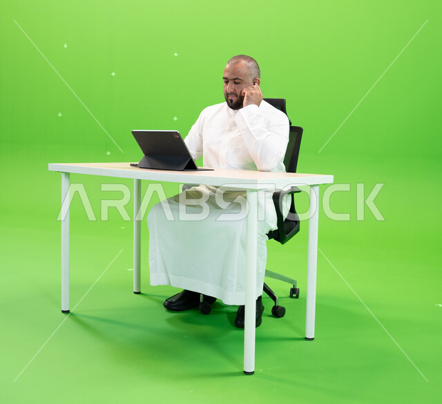 شركات ووظائف سعودية, إنجاز مهام العمل باستخدام جهاز التابليت, العمل في بيئة مريحة, تحقيق أهداف واستراتيجية الشركة، مفهوم دمج العمل مع التقنية والتكنولوجيا, بورتريه لرجل عربي سعودي خليجي يرتدي الثوب السعودي يتحدث عبر سماعة البلوتوث، خلفية خضراء, كروما.