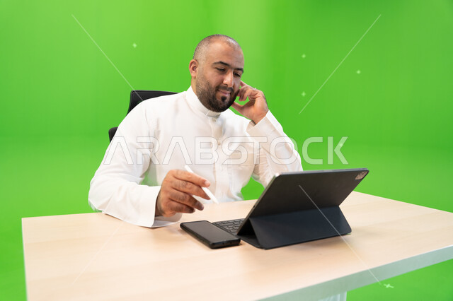 Completing work tasks using a tablet device, working in a comfortable environment, achieving the company’s goals and strategy, the concept of integrating work with technology and technology, a close-up portrait of a Saudi Gulf Arab man wearing the Saudi t