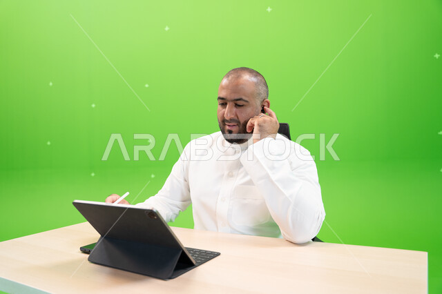 Completing work tasks using a tablet device, working in a comfortable environment, achieving the company’s goals and strategy, the concept of integrating work with technology and technology, a close-up portrait of a Saudi Gulf Arab man wearing the Saudi t