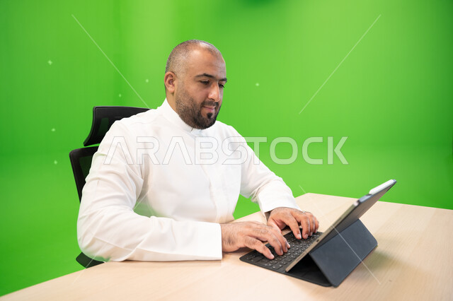 العمل في بيئة مريحة, تحقيق أهداف واستراتيجية الشركة، مفهوم دمج العمل مع التقنية والتكنولوجيا, بورتريه مقرب لرجل عربي سعودي خليجي يرتدي الثوب السعودي التقليدي ينجز مهام العمل باستخدام جهاز التابليت, شركات ووظائف سعودية, خلفية خضراء, كروما.
